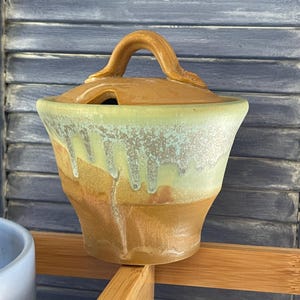 May include: A ceramic sugar bowl with a lid featuring a handle. The bowl has a tan base with a light green glaze dripping down from the rim. The lid is a solid tan color with a curved handle and a small notch for a spoon to rest inside.