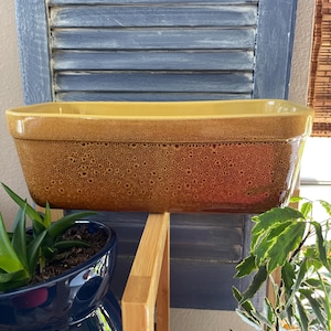 May include: A rectangular, ceramic baking dish with a speckled brown exterior and a yellow interior. The dish is displayed on a wooden shelf against a blue shutter background. The dish is empty and ready for use.