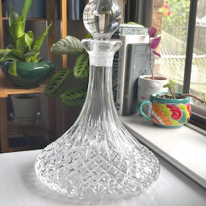 May include: A clear glass decanter with a wide, rounded base featuring a diamond-cut pattern and a smooth, tapered neck. The decanter is topped with a spherical glass stopper that fits into the neck, creating a classic silhouette for serving.