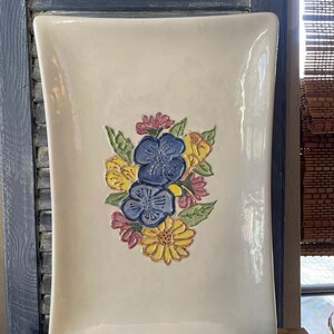 May include: A rectangular ceramic tray with a glossy off-white finish. The center features a raised, hand-painted floral arrangement with two large blue flowers, two yellow daisies, and smaller pink blossoms surrounded by green leaves.