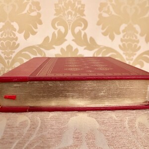 May include: A red hardcover book with gold-colored edges and a decorative pattern on the cover. The book is closed and lying on a patterned surface.