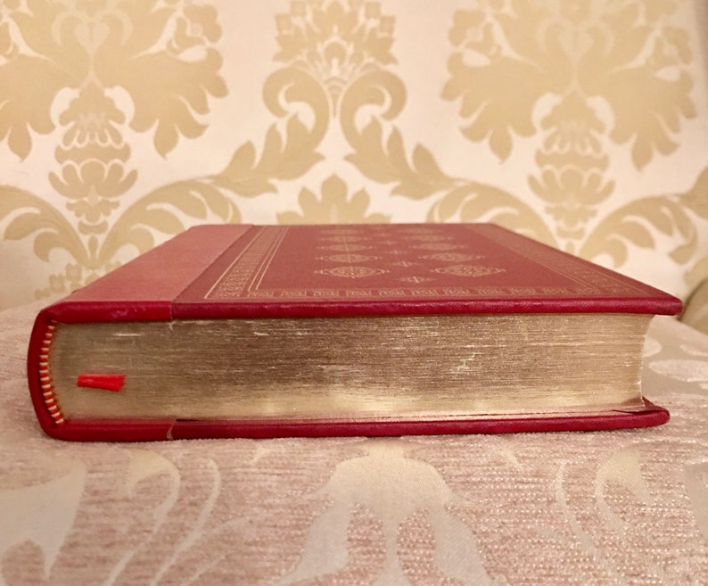 May include: A red hardcover book with gold-colored edges and a decorative pattern on the cover. The book is closed and lying on a patterned surface.