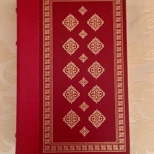 May include: A red hardcover book with a gold border and decorative pattern. The book has a spine with a red ribbon.