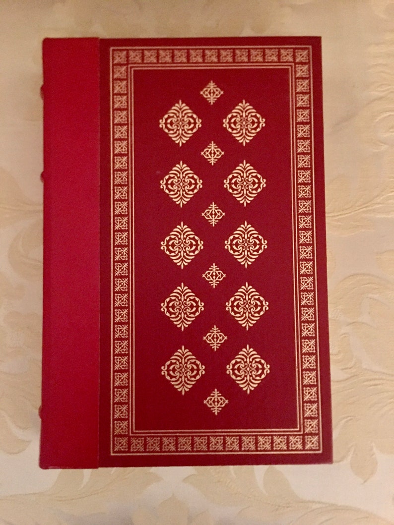 May include: A red hardcover book with a gold border and decorative pattern. The book has a spine with a red ribbon.