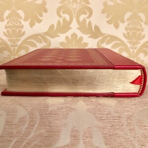 May include: The spine of a red hardcover book with gold accents and a red ribbon bookmark. The book is on a cream-colored surface.