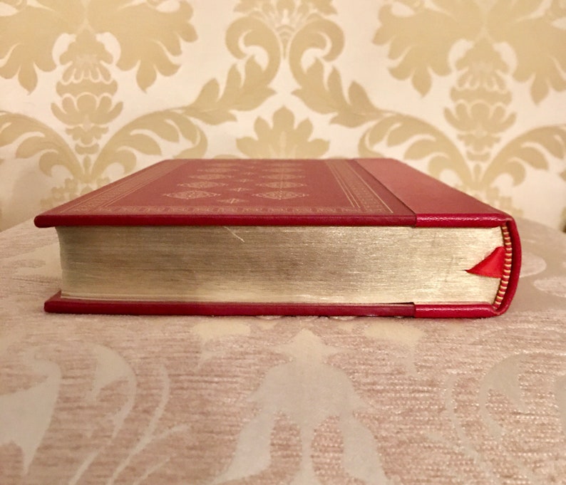May include: The spine of a red hardcover book with gold accents and a red ribbon bookmark. The book is on a cream-colored surface.