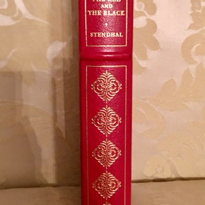 May include: A red hardcover book with gold lettering and decorative accents. The title is "The Red and the Black" by Stendhal.