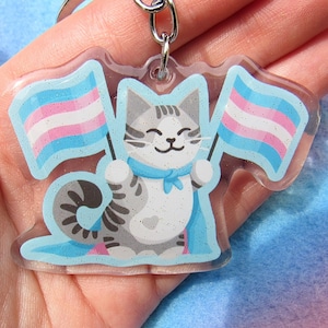Puede incluir: Un llavero acrílico transparente con un gato atigrado gris sonriente sosteniendo dos banderas del orgullo transgénero. El gato tiene una capa y un lazo azules. El llavero tiene un anillo de metal plateado.