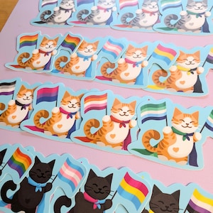May include: A collection of colorful cat stickers with different pride flags. Each sticker features a cartoon cat holding a flag, including the rainbow, transgender, bisexual, and pansexual flags.