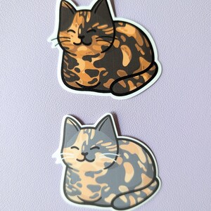 May include: Two calico cat stickers, one with orange and black markings and the other with orange and gray markings. Both cats are smiling and lying down.