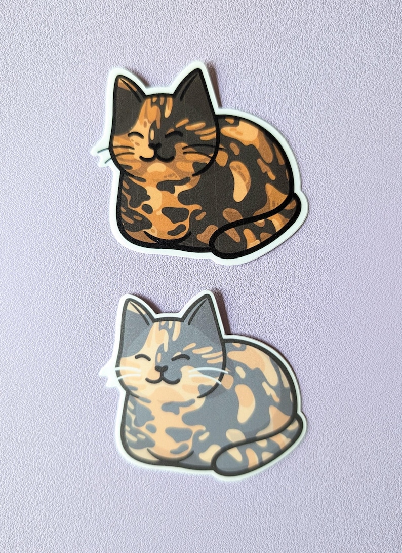 May include: Two calico cat stickers, one with orange and black markings and the other with orange and gray markings. Both cats are smiling and lying down.