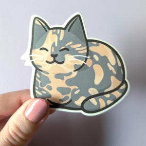 May include: A sticker of a cartoon cat with a calico pattern. The cat is gray, orange, and white with a happy expression. The cat is sitting on its haunches with its tail curled around its body.