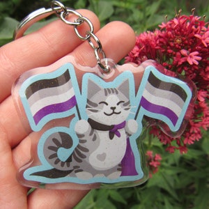 May include: A gray and white cat with a purple cape holds two flags. The flags are the asexual pride flag, with black, gray, white, purple, and blue stripes. The cat is holding the flags in its paws. The keychain is made of clear acrylic and has a silver chain.