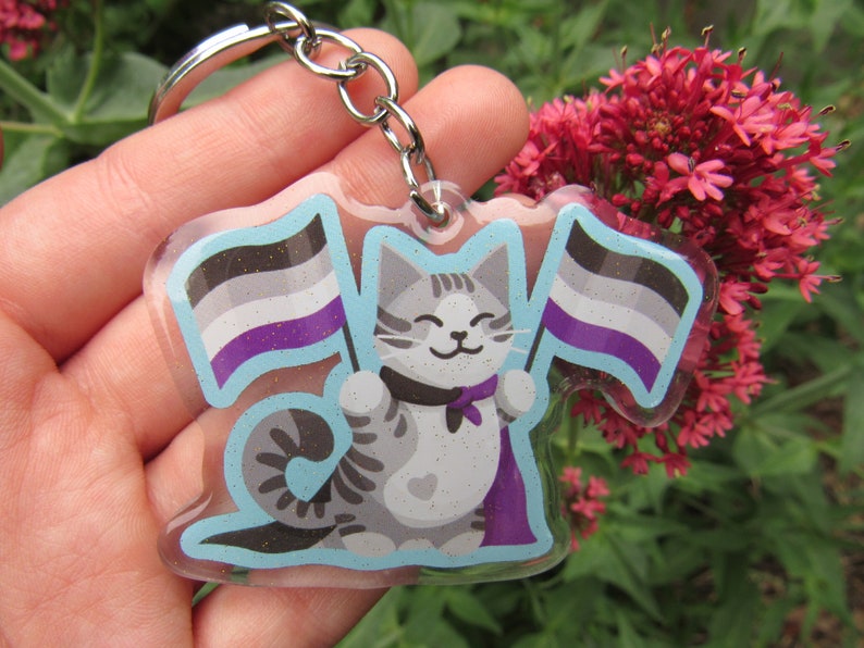 May include: A gray and white cat with a purple cape holds two flags. The flags are the asexual pride flag, with black, gray, white, purple, and blue stripes. The cat is holding the flags in its paws. The keychain is made of clear acrylic and has a silver chain.