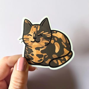May include: A sticker of a cartoon calico cat with orange, black, and white fur. The cat is lying down and smiling.