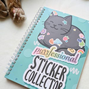May include: A teal blue spiral bound notebook with a black cat illustration. The cat has a rainbow unicorn sticker and other colorful stickers on it. The text "purrfessional sticker collector" is written in rainbow letters on the front of the notebook.