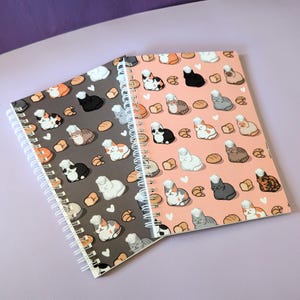May include: Two spiral bound notebooks with a cat and bread pattern. One notebook has a gray cover with white cats and bread. The other notebook has a pink cover with white cats and bread.