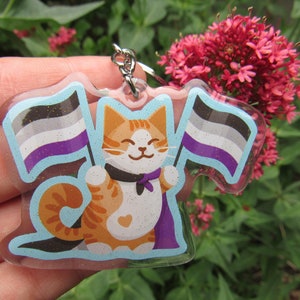 May include: A clear acrylic keychain featuring a cartoon orange tabby cat holding two flags. The flags are the asexual pride flag, with black, gray, white, purple, and blue stripes. The cat is wearing a black bow tie and a purple cape.