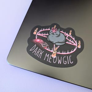 May include: A sticker featuring a gray cat inside a pink pentagram with lit candles. An open book and the words "DARK MEOWGIC" are also included in the design. The sticker is placed on a black laptop.
