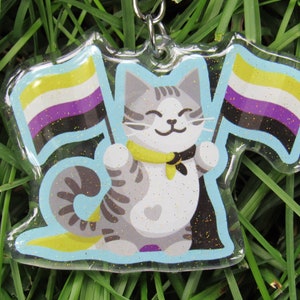 May include: A clear acrylic keychain featuring a cartoon cat holding two flags. The flags are yellow, white, purple, and black, representing the non-binary pride flag. The cat is gray and white with a yellow bow tie. The keychain has a glittery finish.
