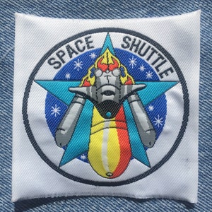 May include: A white patch with a black circle around a blue, yellow and red space shuttle with the text "SPACE SHUTTLE" in black.