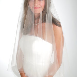 Puede incluir: Una novia con un vestido de novia blanco y un velo blanco. El velo es transparente y cubre su rostro. Ella est&aacute; mirando a la c&aacute;mara con una sonrisa suave.