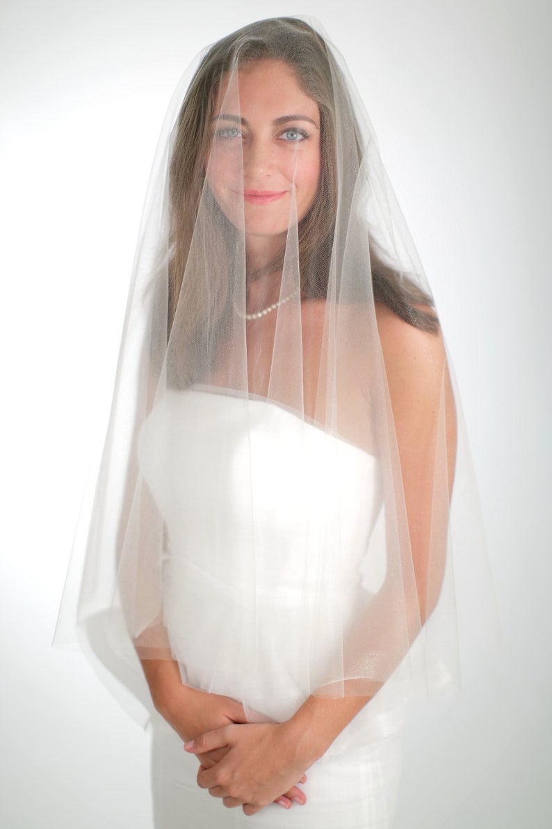 Puede incluir: Una novia con un vestido de novia blanco y un velo blanco. El velo es transparente y cubre su rostro. Ella est&aacute; mirando a la c&aacute;mara con una sonrisa suave.