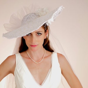 K&ouml;nnte beinhalten: Eine Braut in einem wei&szlig;en Brautkleid mit einem wei&szlig;en Spitzenhut und Schleier. Sie tr&auml;gt eine Perlenkette.