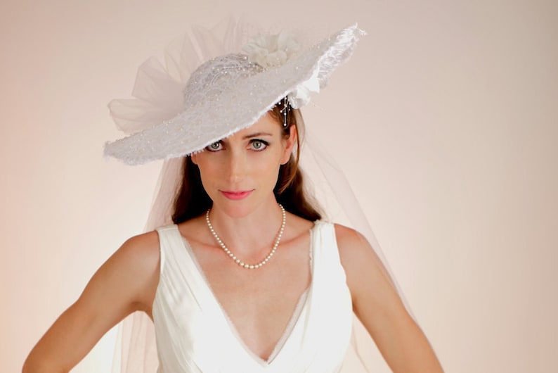 K&ouml;nnte beinhalten: Eine Braut in einem wei&szlig;en Brautkleid mit einem wei&szlig;en Spitzenhut und Schleier. Sie tr&auml;gt eine Perlenkette.