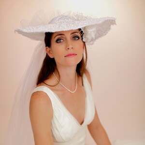 K&ouml;nnte beinhalten: Eine Frau in einem wei&szlig;en Kleid und einem gro&szlig;en wei&szlig;en Hut mit Schleier. Sie tr&auml;gt eine Perlenkette.