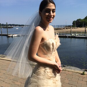 Puede incluir: Una novia con un vestido de novia blanco con velo de tul. El vestido tiene un corpi&ntilde;o ajustado y una falda fluida. La novia est&aacute; de pie en un camino de ladrillos con un cuerpo de agua en el fondo.