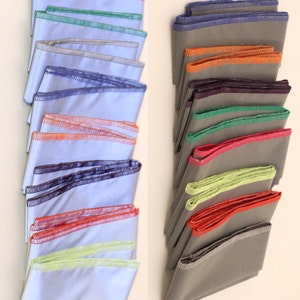 May include: Two stacks of pocket squares. One stack features light blue squares with colourful trim in pink, green, silver, blue, orange, purple, red, and yellow. The other stack has grey squares with trim in orange, purple, green, red, and yellow.