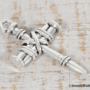 May include: Silver-toned metal cross charm with a nail design. The cross is made of two nails that are intertwined and have a loop at the top for hanging.