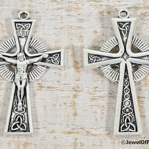 Puede incluir: Dos cruces celtas plateadas con intrincados dise&ntilde;os de nudos. Una cruz presenta una representaci&oacute;n de Jesucristo y las letras INRI. Ambas cruces tienen un lazo en la parte superior para colgar. Las cruces se muestran sobre una superficie clara de vetas de madera.