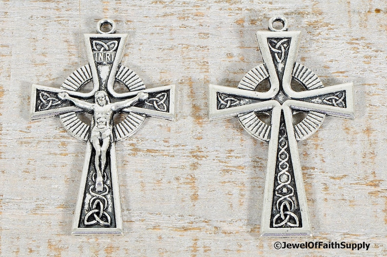 Puede incluir: Dos cruces celtas plateadas con intrincados dise&ntilde;os de nudos. Una cruz presenta una representaci&oacute;n de Jesucristo y las letras INRI. Ambas cruces tienen un lazo en la parte superior para colgar. Las cruces se muestran sobre una superficie clara de vetas de madera.