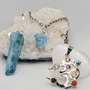 May include: A silver chain necklace with a blue crystal pendant and a silver Om pendant with colorful gemstones. The Om pendant is on a white heart-shaped stone.