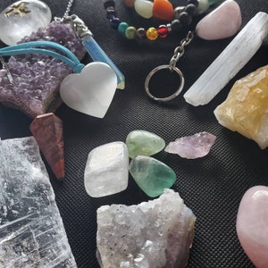 May include: Assortment of crystals and stones, including amethyst, selenite, rose quartz, clear quartz, and green fluorite. The crystals are arranged on a black surface.