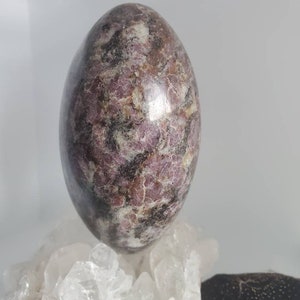 May include: A large, polished, egg-shaped stone with a swirling pattern of pink, white, and black. The stone is resting on a bed of clear quartz crystals.