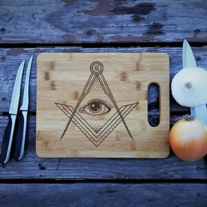 May include: A wooden cutting board with a laser-etched design of an all-seeing eye inside a compass and square. The board has a handle on the side and is made of bamboo.