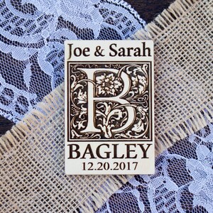 May include: A wooden wedding favour with a decorative initial "B" surrounded by floral designs. The text "Joe & Sarah Bagley 12.20.2017" is printed on the favour.