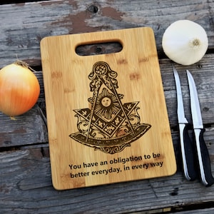 Puede incluir: Una tabla de cortar de madera marrón con un símbolo masónico grabado en ella. La tabla tiene un asa y el texto "You have an obligation to be better everyday, in every way".