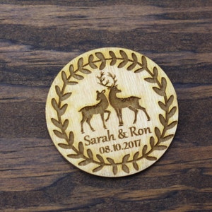 May include: A round wooden wedding favour with a leafy wreath design. Two deer are engraved inside the wreath, and the text "Sarah & Ron 08.10.2017" is below the deer.