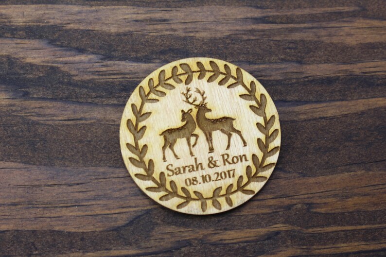 May include: A round wooden wedding favour with a leafy wreath design. Two deer are engraved inside the wreath, and the text "Sarah & Ron 08.10.2017" is below the deer.