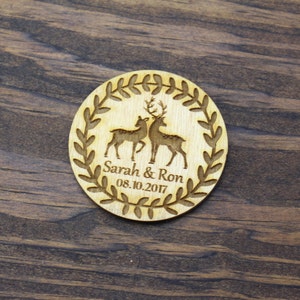 May include: A round wooden wedding favour with a wreath design and two deer silhouettes. The text "Sarah & Ron 08.10.2017" is engraved on the favour.