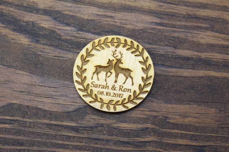 May include: A round wooden wedding favour with a wreath design and two deer silhouettes. The text "Sarah & Ron 08.10.2017" is engraved on the favour.