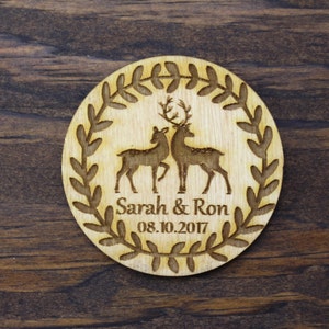 May include: A round wooden coaster with a wreath of leaves around the edge. Inside the wreath are two deer silhouettes, one with antlers. The text "Sarah & Ron 08.10.2017" is printed below the deer.