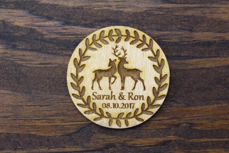 May include: A round wooden coaster with a wreath of leaves around the edge. Inside the wreath are two deer silhouettes, one with antlers. The text "Sarah & Ron 08.10.2017" is printed below the deer.