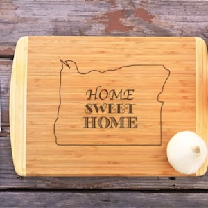 May include: A bamboo chopping board with a wood grain pattern and a natural finish. The board is engraved with the outline of the state of Oregon and the text "Home Sweet Home". There are three knives and three onions on the surface of the board.