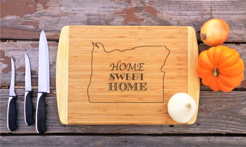 May include: A bamboo chopping board with a wood grain pattern and a natural finish. The board is engraved with the outline of the state of Oregon and the text "Home Sweet Home". There are three knives and three onions on the surface of the board.