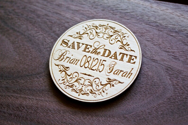 May include: Round wooden save the date sign with a decorative floral design. The sign reads "Save the Date" with the names "Brian & Sarah" and the date "08.12.15".